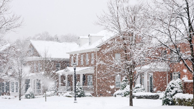 A residential neighborhood during active snowfall showing brick homes with snow-laden trees in the yards and along the street. 